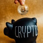 A hand places a Bitcoin into a piggy bank labeled 'Crypto', symbolizing savings and investment.